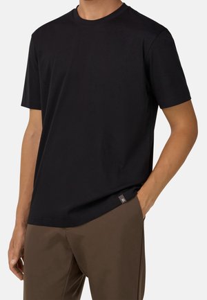 Mężczyzna w jednolitej czarnej koszulce z krótkim rękawem i brązowych spodniach, stojący na tle białego tła z jedną ręką w kieszeni.