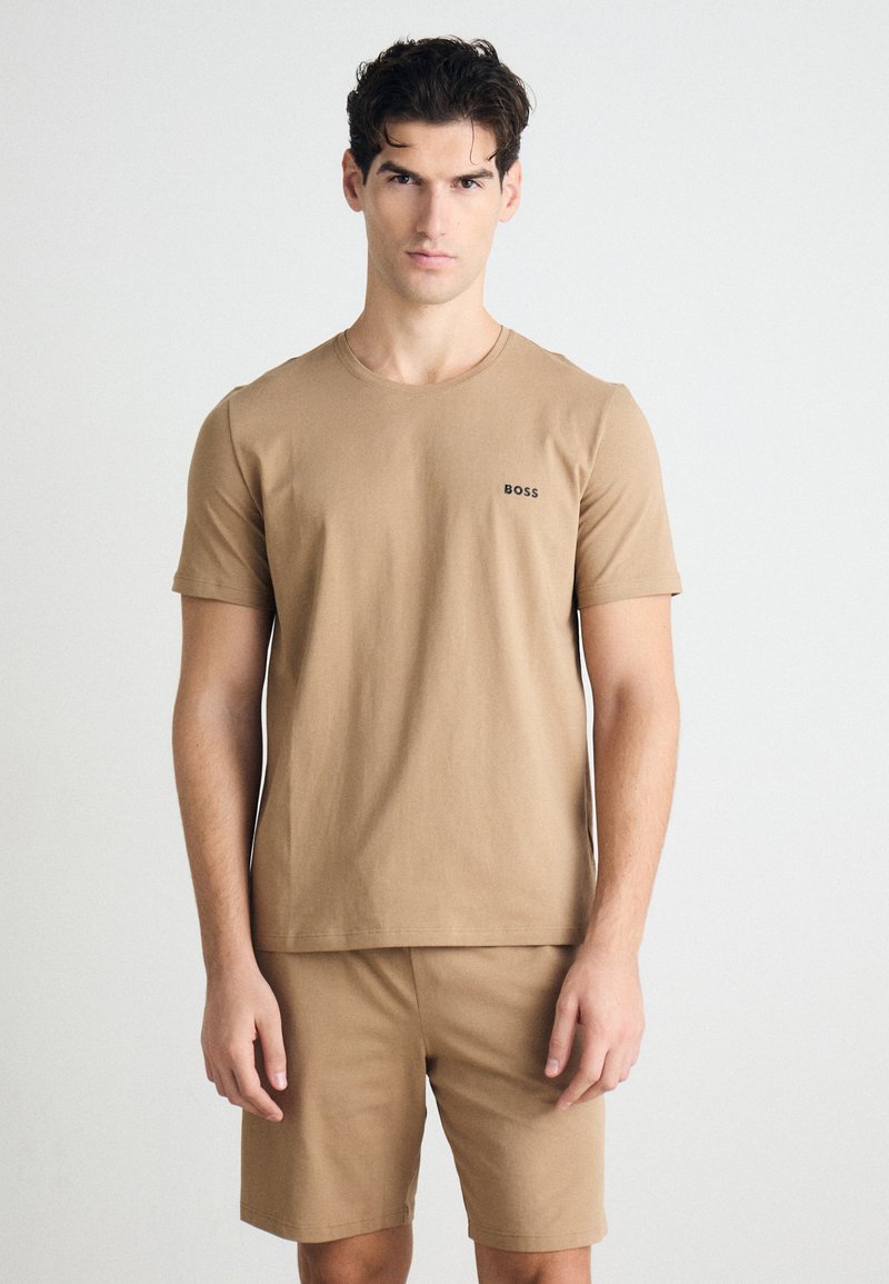 Camiseta de algodón color beige con mangas cortas y cuello redondo, que presenta un logo negro de "BOSS" en el lado izquierdo del pecho; combinada con pantalones cortos a juego.