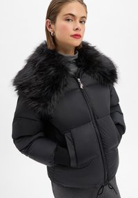 Młoda kobieta w czarnej puchowej kurtce z dużym czarnym futrzanym kołnierzem, szarym golfie i srebrnych kolczykach w kształcie kół, z rękami w kieszeniach.