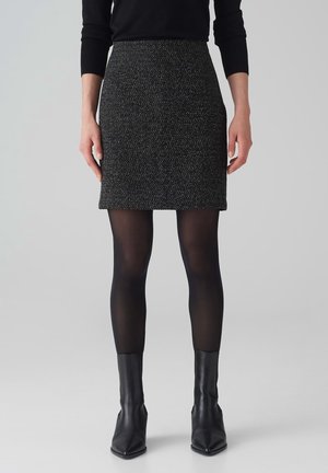 Woman wearing a black textured mini skirt, black sheer tights, black ankle boots, and a black long-sleeve top, standing against a plain background.