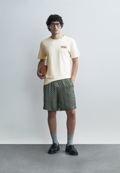 Νεαρός άνδρας με σγουρά μαλλιά και γυαλιά, που φοράει μπεζ μπλουζάκι, πράσινα σορτς με σχέδιο, γκρι κάλτσες, μαύρα παπούτσια και κρατάει ένα καφέ τσαντάκι.