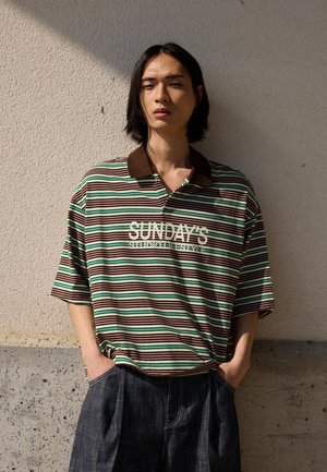 Young person with shoulder-length dark hair wearing a brown, green, and white striped polo shirt with "SUNDAY'S" text, standing against a textured wall.