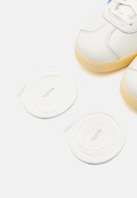 Zapatillas de piel blanca con suela de goma, con acentos en marrón claro, al lado de dos cordones blancos enroscados. Textura suave, diseño minimalista.