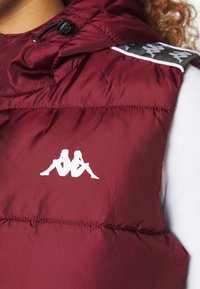 Bordowy, pikowany kamizelka z zamkiem błyskawicznym i kapturem ze sznurkiem. Posiada białe logo na klatce piersiowej oraz wzorzysty akcent w okolicy ramienia. Gładka tekstura.