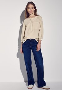 Camicia a quadri beige con maniche tre quarti, abbinata a jeans a gamba larga blu scuro e sneakers bianche. Modella in piedi contro uno sfondo semplice.