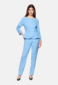 Hellblaues, tailliertes Peplum-Top mit 3/4-Ärmeln, kombiniert mit passenden schmalen Hosen und akzentuiert durch schwarze Absatzschuhe. Glattes, mattes Gewebe.