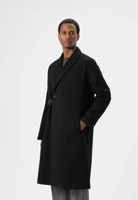 Schwarzer Wollmantel mit Reverskragen, seitlichen Taschen und silbernen Schnallendetails. Figurbetontes Design und mittellange Schnittführung. Glatte Textur.