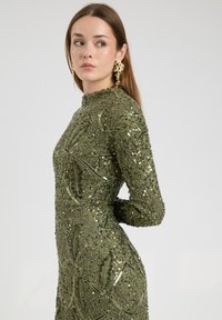 Abito a maniche lunghe di colore verde oliva con intricati motivi di paillettes e ricami. Collo alto, design aderente con dettagli testurizzati.