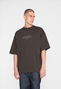 Mørkebrun oversized t-shirt med korte ærmer, rund hals og en grå logo detalje på brystet. Bæres med blå denim bukser.
