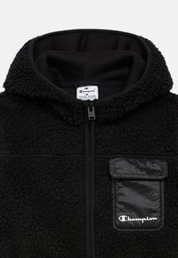 Juodas tekstūruotas fliso džemperis su gobtuvu, priekinis užtrauktukas ir blizgus juodas krūtinės kišenė su balta "Champion" logotipu.
