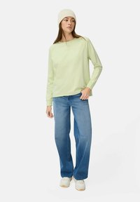 Lichtgroen langemouwen shirt met ronde hals, gecombineerd met losse blauwe jeans. Het model draagt een crèmekleurige beanie en witte sneakers.