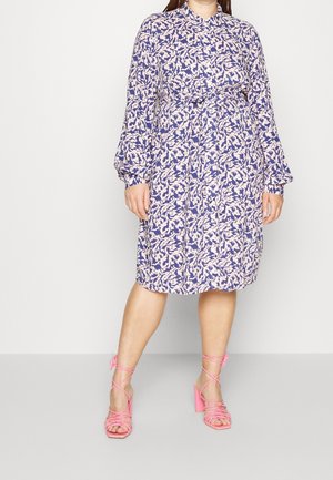 Lila und cremefarbenes Blumenkleid mit langen Ärmeln und einem Bindegürtel, kombiniert mit pinken, geschnürten Sandalen mit Absatz.