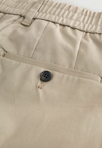 Beige Stoffshorts mit elastischem Bund, einem Knopfverschluss und einer einzelnen Gesäßtasche mit Knopfdetails. Glatte Textur.