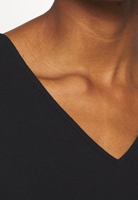 Haut noir à col en V avec une texture lisse, des coutures précisément cousues et une coupe ajustée mettant en valeur la zone de la clavicule.