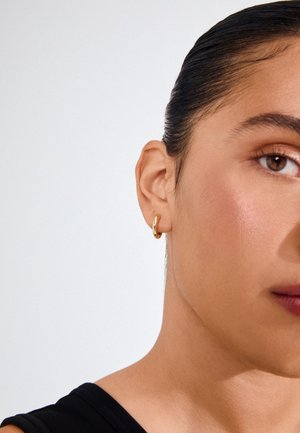 Kuldne rõngas kõrvarõngas inimese kõrvus, millel on sile ja poleeritud viimistlus. Taust on lihtne valge, rõhutades kõrvarõnga disaini.