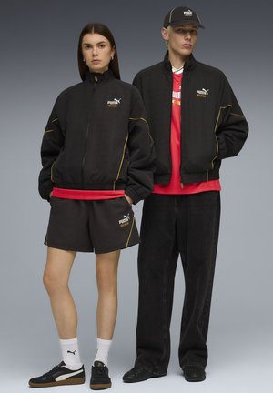 Veste noire zippée avec accents jaunes, chemise rouge, shorts noirs et baskets. Le modèle se tient devant un fond gris, présentant une tenue sportive.