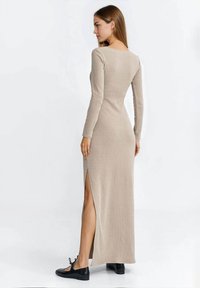 Długa beżowa prążkowana sukienka z długimi rękawami, rozcięciami po bokach, dopasowanym krojem i gładką fakturą, zestawiona z czarnymi płaskimi butami.