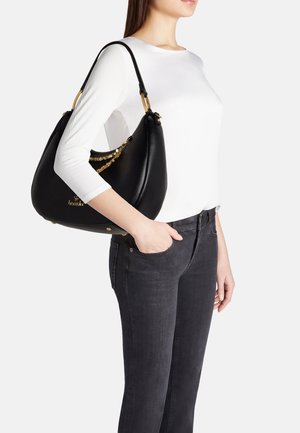 Vrouw draagt een wit shirt met lange mouwen en zwarte spijkerbroek, met een zwarte leren handtas met gouden kettingdetail op haar schouder.