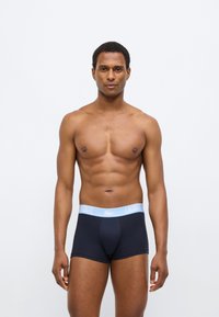 Boxer bleu marine avec une ceinture élastique bleu clair arborant le logo Lacoste, porté par un modèle masculin. Tissu lisse, coupe ajustée.