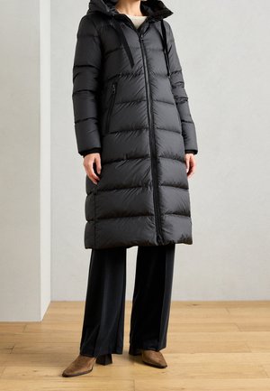 Person iført en lang sort quiltet dunjakke med hætte, sorte bukser med vide ben og brune spidse hælede støvler står på et trægulv.