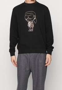 Felpa nera con grafica di un personaggio dei cartoni animati che si illumina in bianco e oro, con scollatura e polsini a costine. Abbinata a pantaloni grigi.