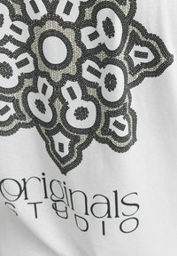 Bílá látka s geometrickým potiskem mandaly v černé a olivově zelené barvě. Pod textem je napsáno "originals studio" moderním písmem.