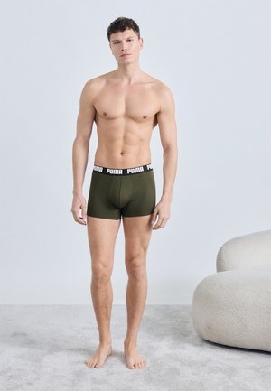 Jeune homme debout pieds nus sur un tapis, portant un boxer Puma vert olive, à côté d'un fauteuil incurvé de couleur crème.