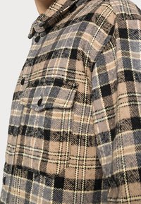 Brun och grå rutigt skjorta i flingor, med krage, två svarta knäppfickor och ett strukturerat tyg med mönster i flera riktningar.