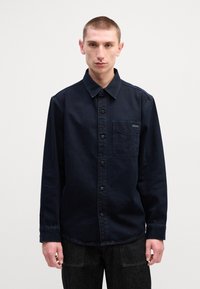 Camisa de ganga azul-escura com fecho de botão à frente, um único bolso no peito e colarinho pontudo. Apresenta mangas compridas e um corte descontraído.