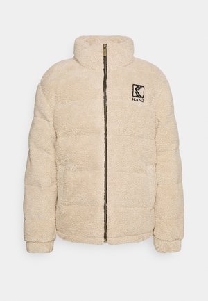 Beige fleece jakk, millel on kõrge krae, tõmblukuga kinnitamine, polsterdatud disain ja rinna peal broddeeritud logo. Pehme, tekstuuriga välimus.