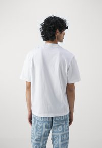 White cotton t-shirt with a crew neck, relaxed fit. Paired with light blue jeans featuring repeating bold text patterns.