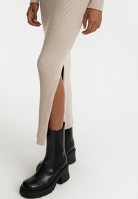 Geribbelde, lichtbeige gebreide jurk met een zijopening tot boven de dij, gecombineerd met chunky zwarte leren enkelboots met een dikke zool en korte schacht.