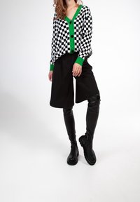 Cárdigan a cuadros en blanco y negro con acentos verdes, combinado con pantalones cortos negros y botas altas negras con un acabado texturizado.