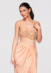 Femme aux cheveux foncés portant une robe en satin rose avec un corsage orné et une taille plissée, touchant une épaule et regardant sur le côté sur fond uni.
