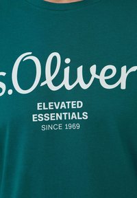 Turkusowa koszulka bawełniana z dużym białym logo z napisem „s.Oliver” oraz „ELEVATED ESSENTIALS” wraz z rokiem „1969” poniżej.