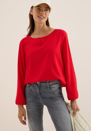 Röd långärmad blus med rund halsringning och nyckelhålsdetalj, tillsammans med figurnära grå jeans. Slät tygkvalitet och avslappnad passform.