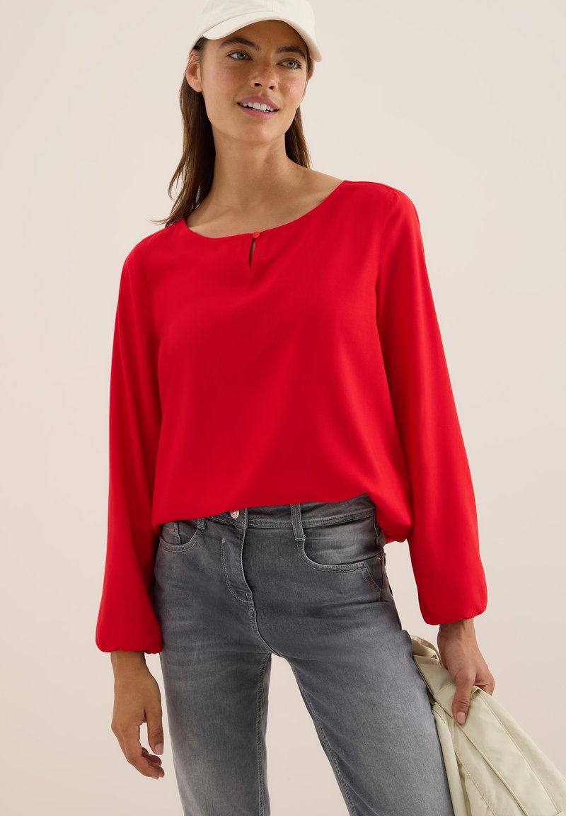 Röd långärmad blus med rund halsringning och nyckelhålsdetalj, tillsammans med figurnära grå jeans. Slät tygkvalitet och avslappnad passform.