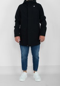 Veste noire imperméable avec capuche, dotée d'une fermeture à l'avant, de deux poches latérales et d'un petit logo blanc. Portée sur un jean en denim bleu et des baskets blanches.