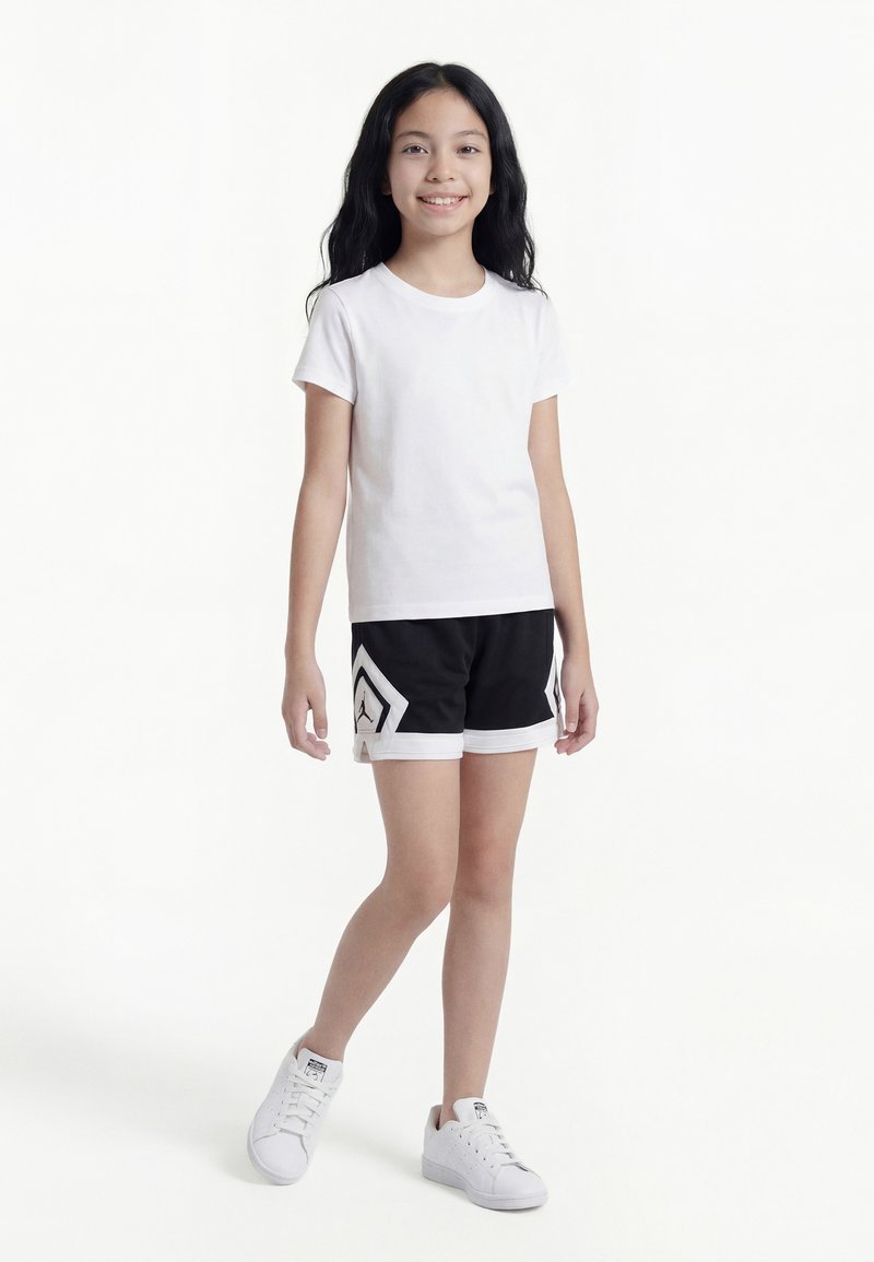 Ragazza giovane in piedi, che indossa una t-shirt bianca, pantaloncini sportivi neri con un logo e scarpe da ginnastica bianche, su sfondo bianco semplice.