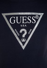 T-shirt blu navy con un design a triangolo metallico argento. Il testo include "GUESS U.S.A.", "WASHED", "JEANS" e un grande punto interrogativo al centro.