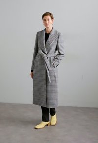 Long gray plaid coat with a tie waist, notch lapels, and large pockets, paired with black trousers and yellow leather shoes.