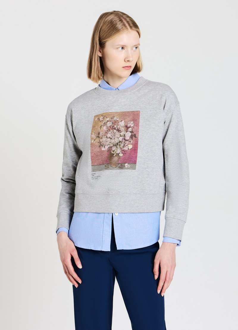 Maglione grigio a maniche corte con una stampa floreale sul davanti, posizionato su uno sfondo pastello. Indossato sopra una camicia colletto azzurra.