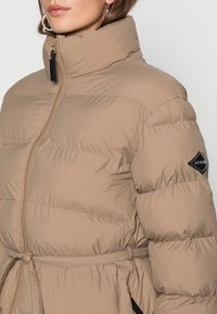 Beige polstret jakke med høj krave, lynlås og bælte i taljen. Har struktureret stof og et sort logo patch på ærmet.