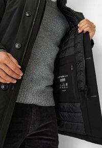 Sort frakke med en quiltet indvendig foring og grå sweater. Har knapper, lynlås og en lomme med plejeanvisninger indvendigt.