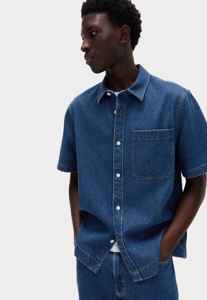 Junger Mann mit dunkler Haut, der ein kurzärmeliges, blaues Jeanshemd mit Knöpfen und eine blaue Jeans trägt, die Hände in den Taschen, blickt zur Seite.
