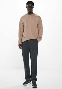 Helle beige Langarmbluse kombiniert mit dunkelgrauen Hosen. Das Model trägt dunkle Sneaker, mit einem einfachen und lockeren Schnitt.