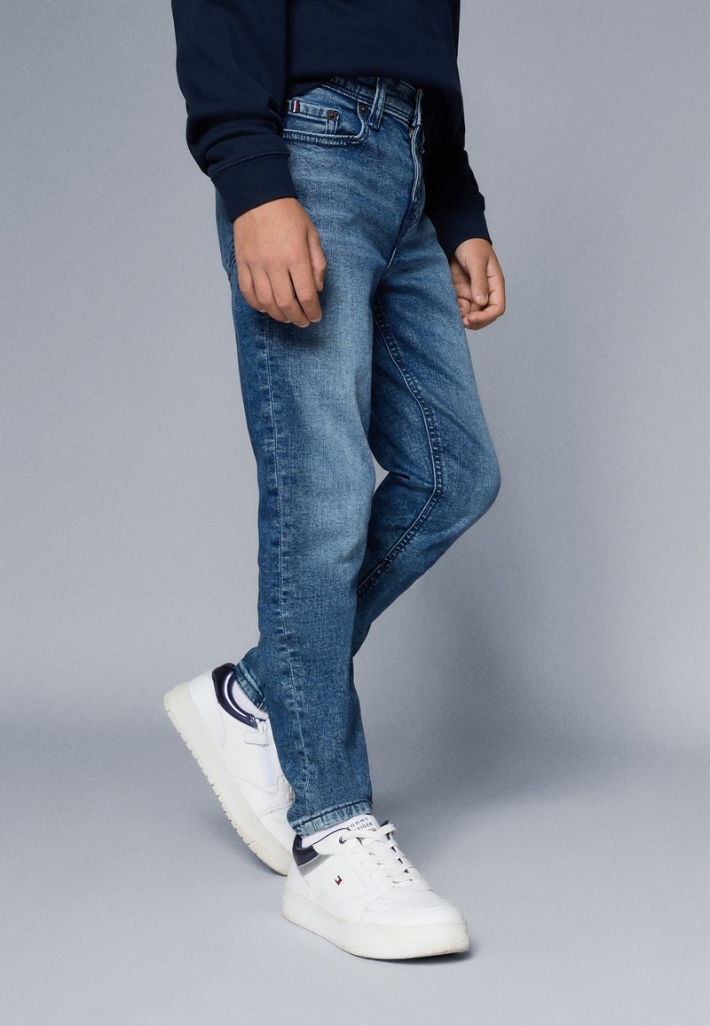 Personne portant un jean bleu, des baskets blanches et un sweat-shirt bleu marine, debout devant un fond gris uni.