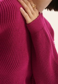 Różowy sweter dziany z teksturowanym wzorem, ściągaczami przy rękawach i luźnym krojem, z ręką opartą na ramieniu, prezentującą złoty pierścionek.