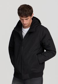 Giacca nera con zip e cappuccio, caratterizzata da un'estremità liscia, polsini e orlo a coste, e due tasche laterali. Indossata con una t-shirt bianca sotto.