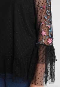 Haut à pois noirs avec des manches évasées transparentes ornées de broderies florales colorées. Ourlet volanté et tissu texturé.